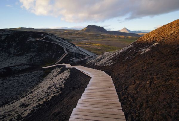 Holz-Laufsteg durch eine schroffe, bergige Landschaft, weiter Blick über eine grüne Ebene