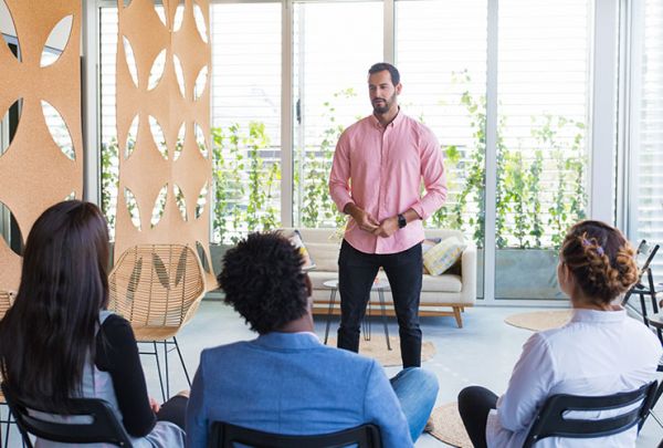 ein Mann steht vor 3 Zuhörenden die sitzen, hinter ihm ist ein großes Fenster, er trägt ein rosafarbenenes Hemd und spricht zum Thema Achtsamkeit