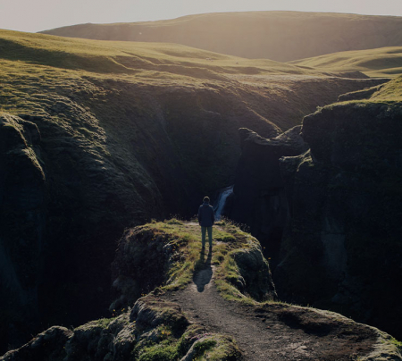 Mann steht auf einem Berg an einer Klippe, vor ihm eine Schlucht und ein Wasserfall, der Mann schaut weg vom Betrachter