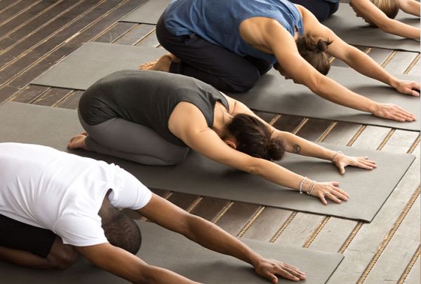 4 Personen, ein schwarzer Mann, drei weiße Frauen, praktizieren Yoga in der Balasana-Stellung. Mittelgraue Yogamatten, Holz-beplankter Fussboden